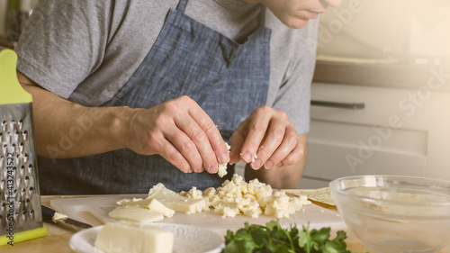 A man without a face in an apron in the kitchen prepares a dish of cheese, cuts it with his hands on a board, and grates it. Homemade Cheese Cooking Process, Culinary Homemade Hobby