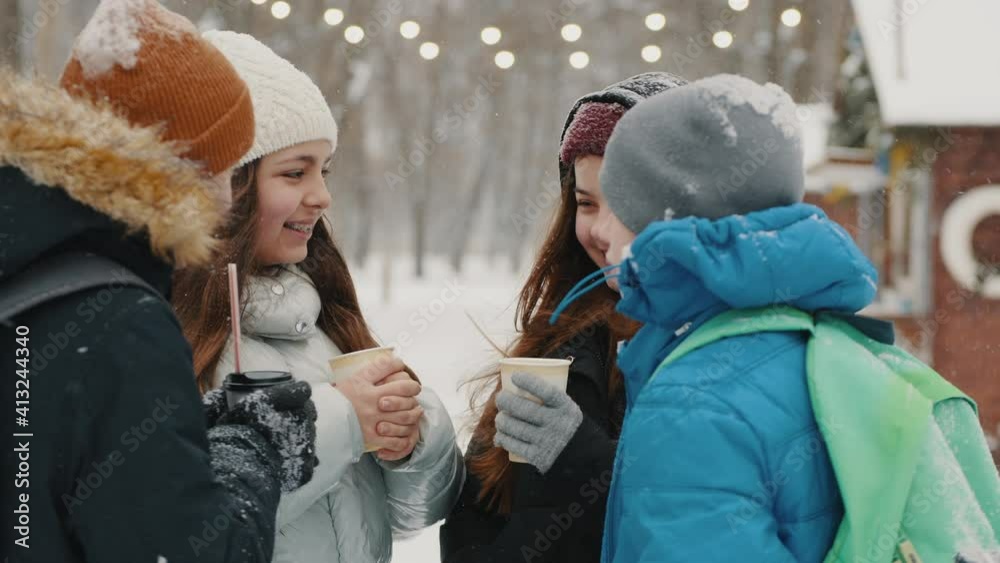 Teenagers spend time together on a walk in the park in winter in snowy weather, classmates smile, talking and drink warm drinks from paper cups Teenagers spend time together on a walk in the park in winter in snowy weather, classmates smile, talking and drink warm drinks from paper cups