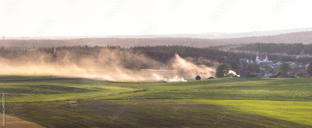 Fototapeta premium Panorama of the countryside of Belarus. Agricultural fields view. Fresh green field and a small European town. Smoke from burnt grass spreads across the field. Large forest in fog on the background