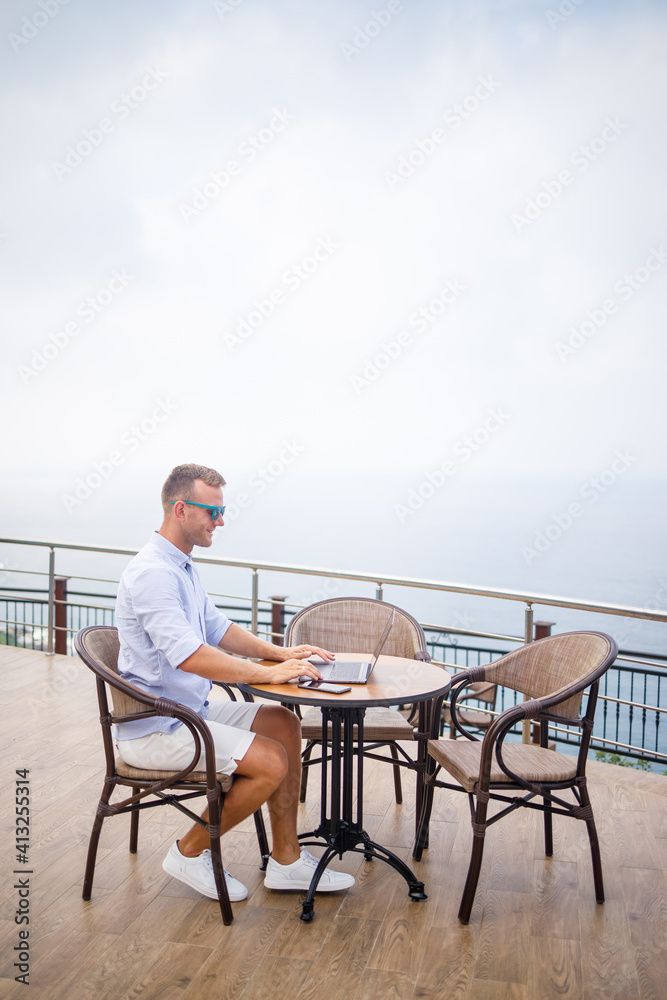 Handsome successful young male businessman sitting at a table by the pool with a laptop overlooking the Mediterranean Sea. Remote work on vacation. Vacation concept