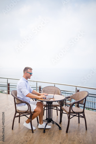 Handsome successful young male businessman sitting at a table by the pool with a laptop overlooking the Mediterranean Sea. Remote work on vacation. Vacation concept © Дмитрий Ткачук