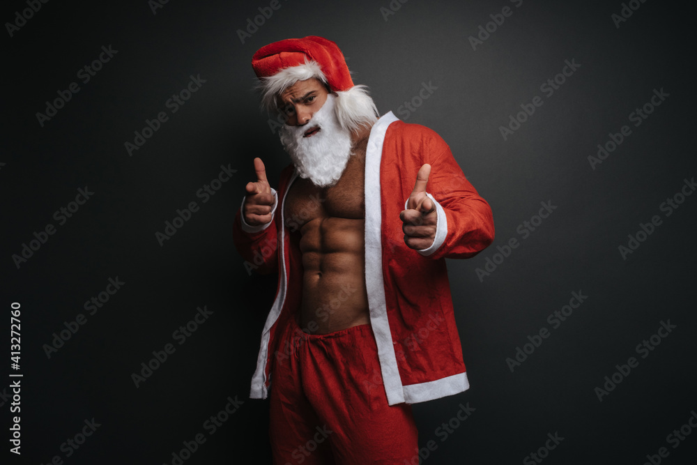 Pointing fingers at camera muscular man in Santa Claus costume in studio. Emotional portrait of handsome male model in Christmas outfit at grey background. Pointing fingers at camera muscular man in Santa Claus costume in studio. Emotional portrait of handsome male model in Christmas outfit at grey background.