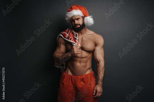 Sad Santa Claus with red plaid in studio. Sexy muscular guy in Santa hat at grey background. Shirtless man in Christmas pants holding  checkered blanket on his chest.