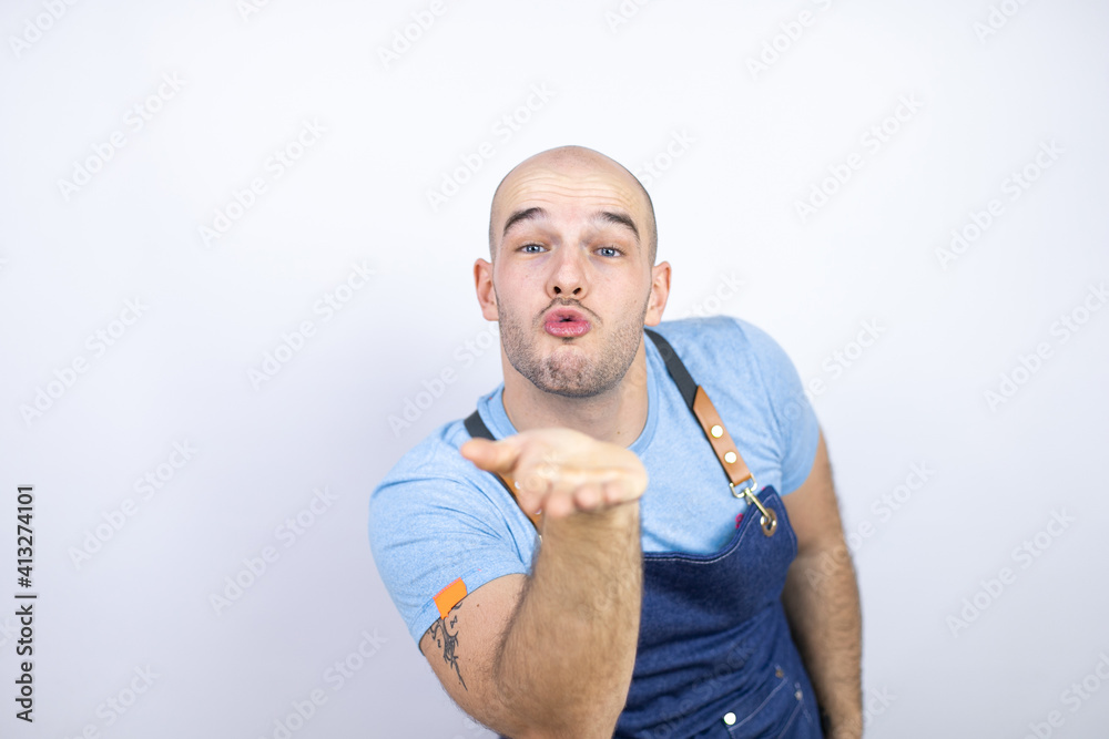 Young bald man wearing apron uniform over isolated white background looking at the camera blowing a kiss with hand on air being lovely and sexy. Love expression. Young bald man wearing apron uniform over isolated white background looking at the camera blowing a kiss with hand on air being lovely and sexy. Love expression.