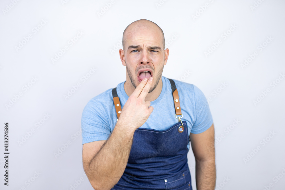 Young bald man wearing apron uniform over isolated white background disgusted with his hand inside his mouth Young bald man wearing apron uniform over isolated white background disgusted with his hand inside his mouth