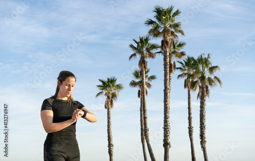 Fitness young female looking smart watch after running with beautiful palm trees on background.Sports technology and healthy lifestyle concept with copy space.