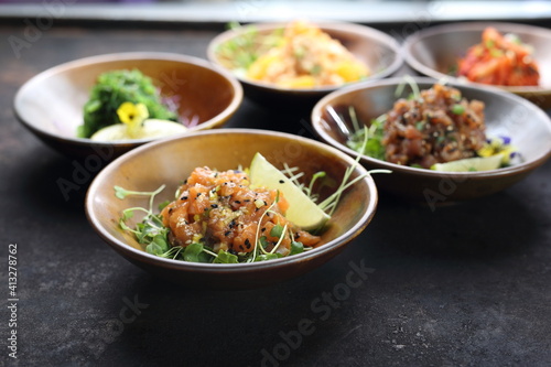 A composition of appetizers: tuna tartare, salmon tartare, seaweed salad, kimchi, shrimps salad, on a black stony background.