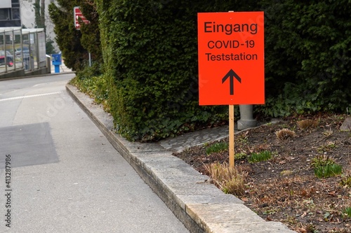 Test station Covid-19 signpost in German in orange color showing with arrow the direction to the testing center on the ground of Zurich University Hospital in Switzerland. It is beside sidewalk.