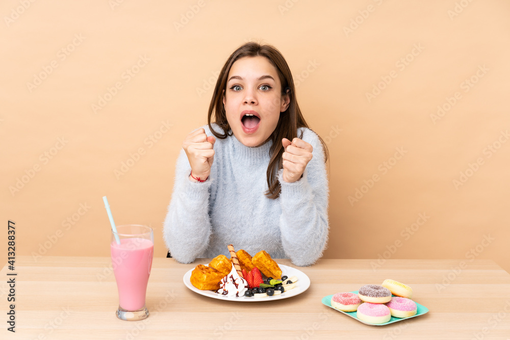 Teenager girl eating waffles isolated on beige background celebrating a victory in winner position Teenager girl eating waffles isolated on beige background celebrating a victory in winner position