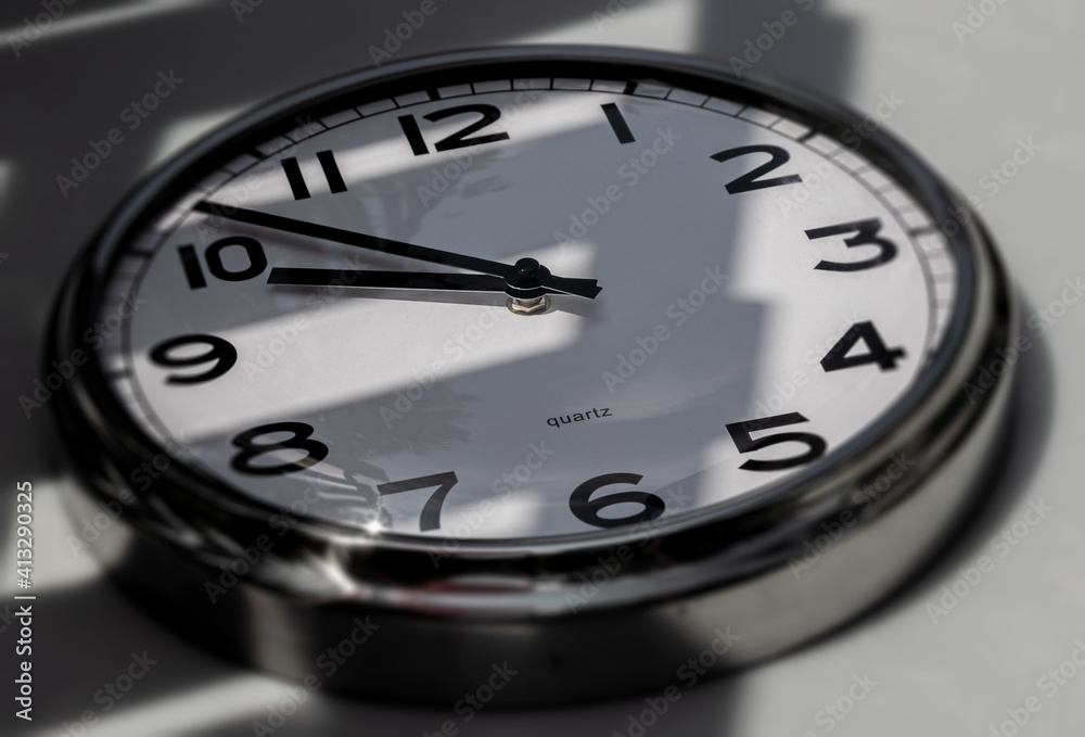 A Modern white aluminum wall clock in sunlight with shadows on the clock face. Close up to a wall clock. Reflections in the aluminum frame. The pointers at 10 to 10. A Modern white aluminum wall clock in sunlight with shadows on the clock face. Close up to a wall clock. Reflections in the aluminum frame. The pointers at 10 to 10.