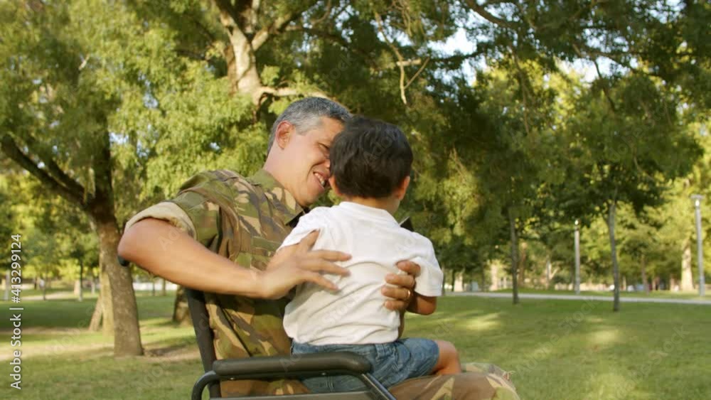 Disabled military dad walking and playing with kids in park, holding boy in arms on lap, while girl pushing wheelchair. Static shot. Veteran of war or returning home concept Disabled military dad walking and playing with kids in park, holding boy in arms on lap, while girl pushing wheelchair. Static shot. Veteran of war or returning home concept