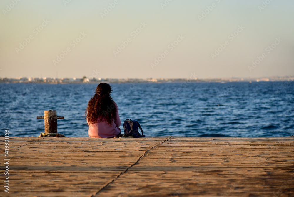 A young girl sits on a sea pier and is sad, view from the back.