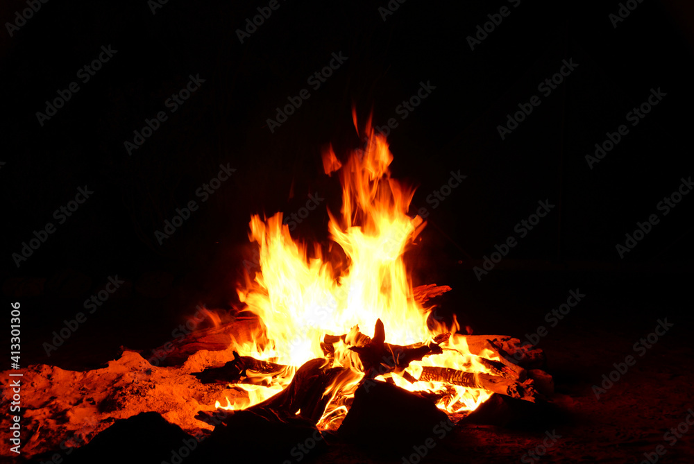 Bright flames, embers, and burning wood illuminating the ground around a camp fire.