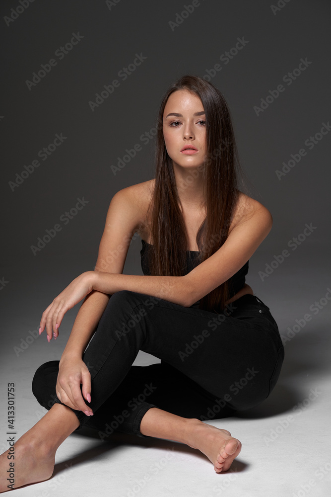 Full length studio portrait of young slim tanned caucasian girl in black jeans and bando top sitting and posing against grey studio background