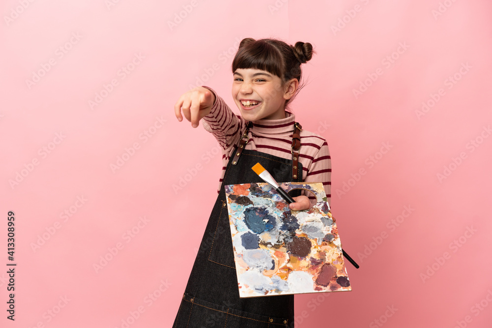 Little artist girl holding a palette isolated on pink background pointing front with happy expression Little artist girl holding a palette isolated on pink background pointing front with happy expression