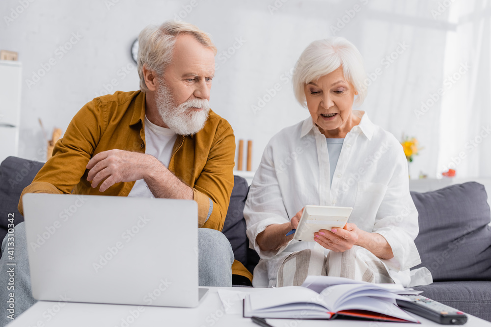 Senior woman using calculator near husband, laptop and notebook on blurred foreground Senior woman using calculator near husband, laptop and notebook on blurred foreground