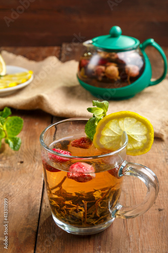 green tea in a glass cup with strawberries mint and lemon on a wooden table and a teapot and lemon in a plate and mint leaves.
