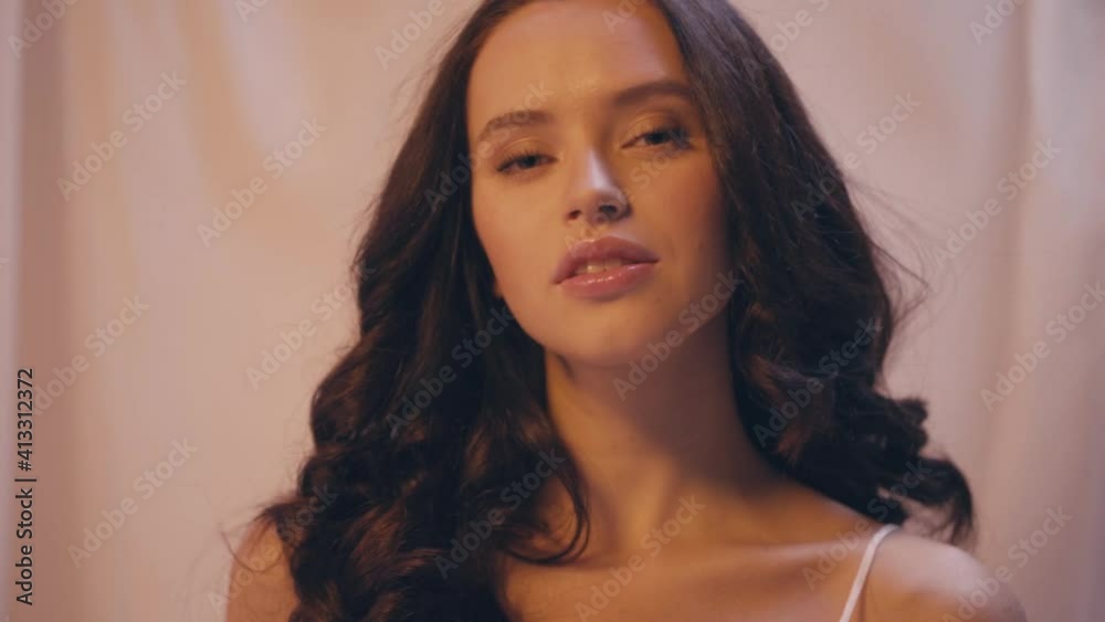 slow motion of sensual woman adjusting wavy hair and looking at camera on white