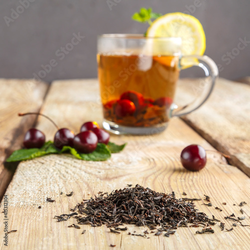 Fototapeta Naklejka Na Ścianę i Meble -  black tea in a glass cup with mint cherries and lemon on a wooden table next to fresh cherries and sprinkled tea leaves.