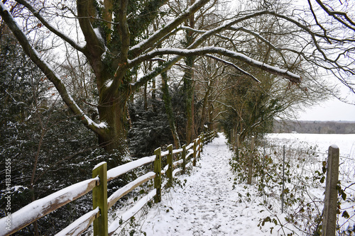 View looking down a side path of the Worth Way near the village of Crawley Down in West Sussex, England, UK. Snowy footprints cover the path with snow covered tree branches and wooden fencing.
