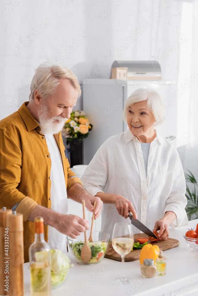 Senior man mixing salad near smiling wife and wine on blurred foreground Senior man mixing salad near smiling wife and wine on blurred foreground