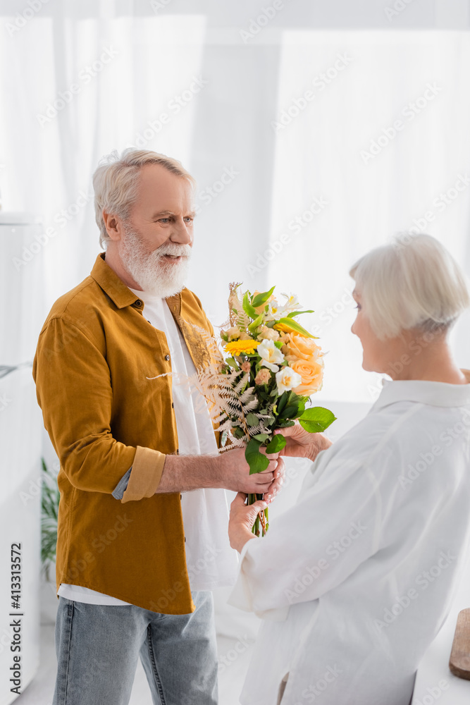 Smiling senior man giving bouquet to wife on blurred foreground in kitchen Smiling senior man giving bouquet to wife on blurred foreground in kitchen