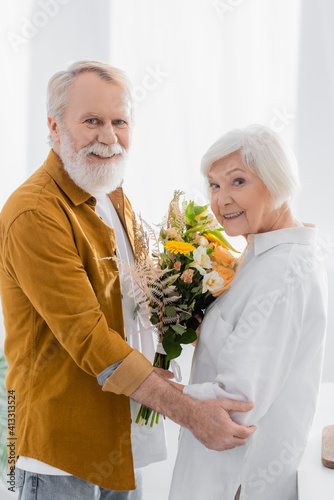 Senior couple with flowers smiling at camera in kitchen