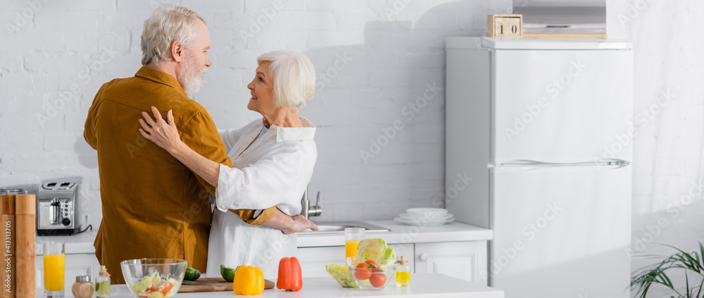 Positive elderly couple hugging near ripe vegetables on kitchen table, banner - obrazy, fototapety, plakaty 