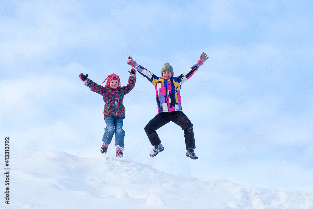 Winter activities outdoors. Happy expressive little girls wearing a warm clothes jumping and enjoying life against the blue sky on a snow in winter Winter activities outdoors. Happy expressive little girls wearing a warm clothes jumping and enjoying life against the blue sky on a snow in winter