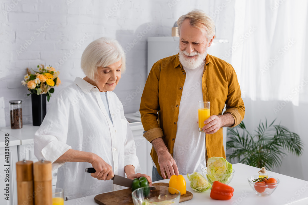 Senior man holding glass of orange juice near wife cutting paprika while cooking in kitchen