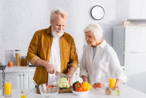 Senior man cutting cabbage near vegetables and wife with orange juice