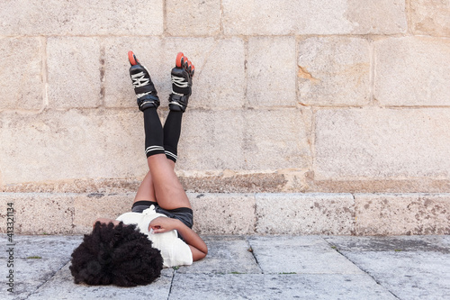 Young African American woman with afro hair is getting restless lying on the floor with her legs up with inline skates for training outdoor skating and playing sports on a hot summer day