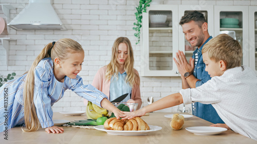 Children taking croissants near parents on blurred foreground in kitchen