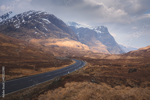 A moody, dramatic mountain landscape of an empty winding A82 road through Glencoe in the Scottish Highlands to the ridges of Beinn Fhada, Gearr Aonach and Aonach Dubh (Three Sisters of Glencoe).