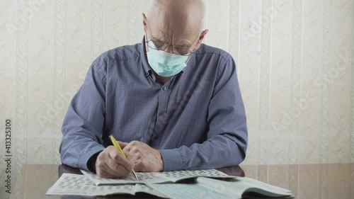 Aged pensioner in medical mask and glasses solves crossword puzzle sitting at table during selfisolation photo