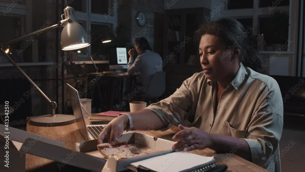 Waist-up shot of african american woman eating pizza while working on laptop staying late in office Waist-up shot of african american woman eating pizza while working on laptop staying late in office