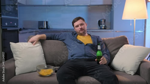 Single unshaven fat man suffering from insomnia switches TV channels on TV lying on couch and drinking alcoholic beer and potato chips on sofa in living room. Man spends evening at home in front TV. photo