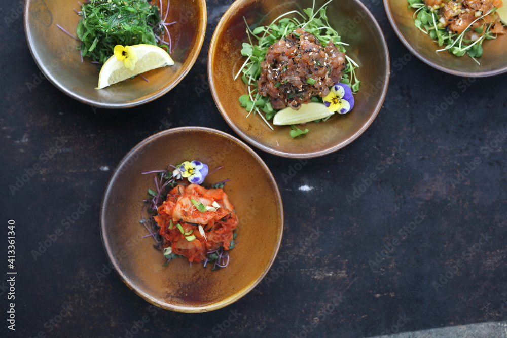 A composition of appetizers: kimchi salad, tuna tartare, seaweed Japanese salad, salmon tartar on a black stony background.