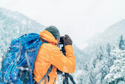 Photographer dressed orange softshell jacket with backpack making a landscape shoot using a digital camera and tripod while he trekking winter mountains route. Photographer occupation concept image.