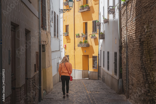 A young blonde female tourist exploring the narrow cobblestone backstreet of the old town (Albaicin or Arab Quarter) in Granada, Andalusia, Spain.