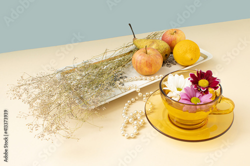 Creative arrangement with cup of tea, colorful blossom,  herbs, and jusy fruit on plate, against pastel  beige background. Minimal concept.