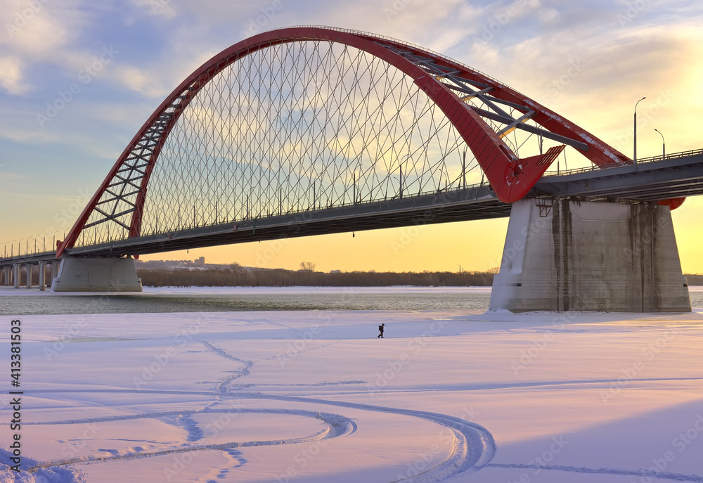 Obraz premium Winter morning on the Ob. A small man under the arch of the Bugrinsky automobile bridge on the bank of a frozen river in Novosibirsk