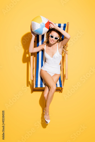 top view of smiling woman in swimsuit sitting in deck chair with inflatable ball on yellow.