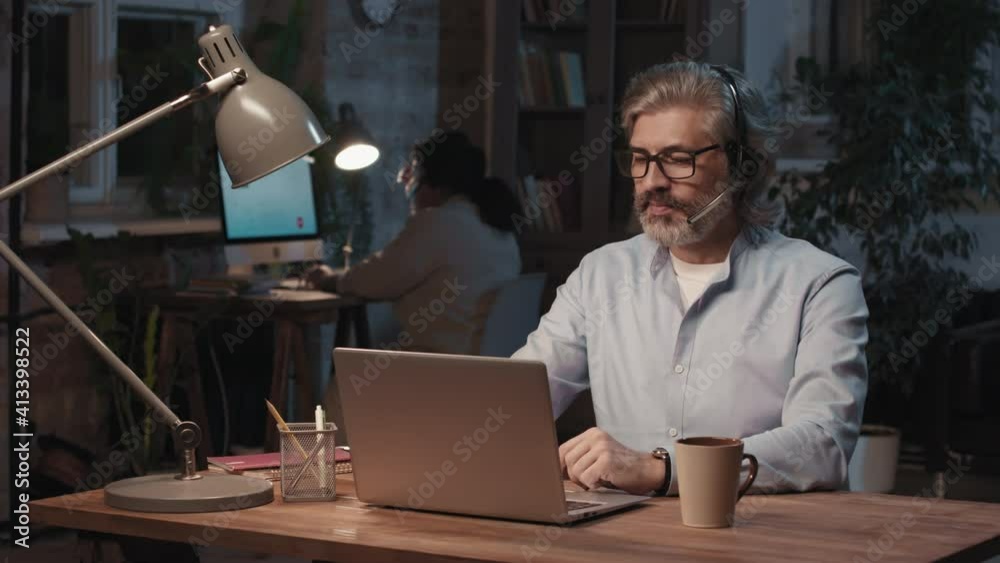 Medium slow-motion portrait of senior caucasian call center operator looking at camera smiling working at nightshift in office Medium slow-motion portrait of senior caucasian call center operator looking at camera smiling working at nightshift in office