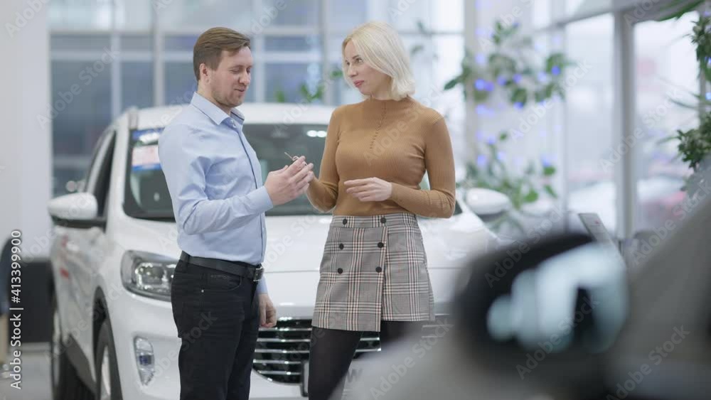 Rich blond slim woman passing car key to happy Caucasian man in showroom. Portrait of successful businesswoman buying new vehicle for husband in car dealership. Gifts and luxury concept.