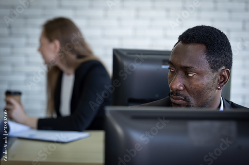 An African man looked around the office with a serious expression. A male office worker paranoid his coworker as he is secretly doing something on the desktop.