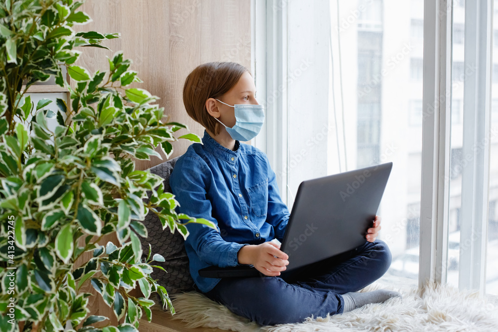 A masked girl is sitting on a windowsill with a laptop. Cozy comfortable learning at home from a distance. He looks thoughtfully out the window. A masked girl is sitting on a windowsill with a laptop. Cozy comfortable learning at home from a distance. He looks thoughtfully out the window.