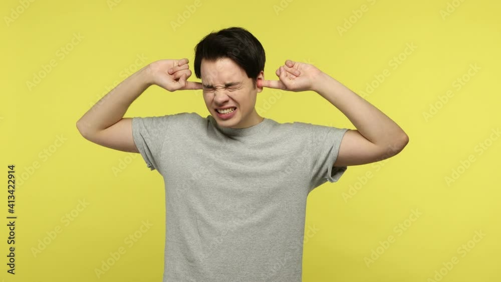 Irritated young man in gray t-shirt closing eyes and ears with fingers and loudly screaming, ignoring conflict, not listening to annoying talk. Indoor studio shot isolated on yellow background Irritated young man in gray t-shirt closing eyes and ears with fingers and loudly screaming, ignoring conflict, not listening to annoying talk. Indoor studio shot isolated on yellow background