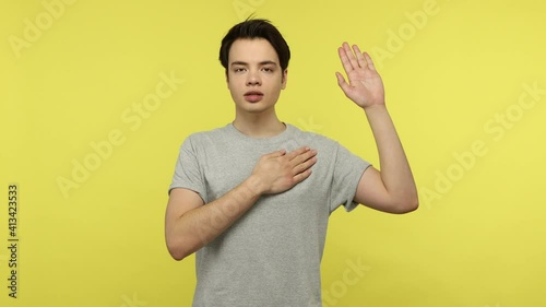Responsible young guy in gray t-shirt making promise holding hand on chest, looking into camera with devoted eyes, swearing fidelity, taking oath. Indoor studio shot isolated on yellow background