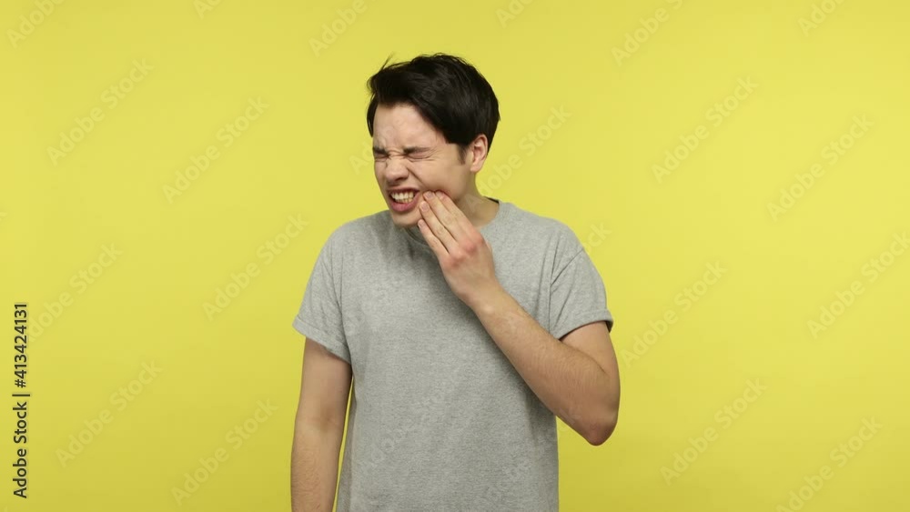 Dental problems. Unhealthy young man in gray t-shirt touching sore cheek frowning from pain, suffering toothache, cavities or gingivitis symptoms. Indoor studio shot isolated on yellow background Dental problems. Unhealthy young man in gray t-shirt touching sore cheek frowning from pain, suffering toothache, cavities or gingivitis symptoms. Indoor studio shot isolated on yellow background
